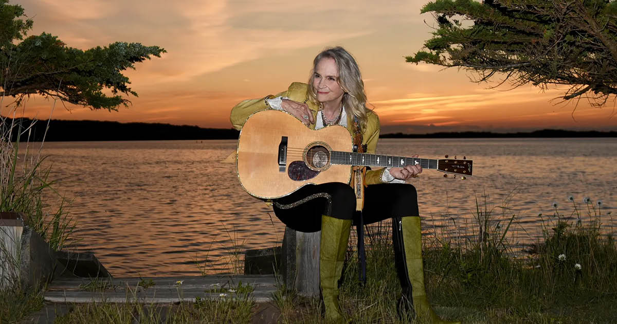 Kate Taylor poses with a guitar on her knee by a lake at sunset